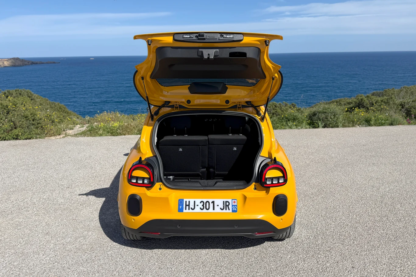 żółty samochód z otwartą klapą bagażnika stoi na utwardzonej nawierzchni, w tle rozciąga się widok na morze i niską roślinność