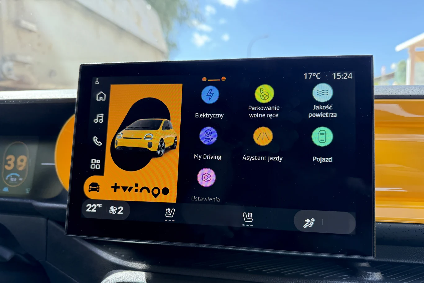 Wyświetlacz samochodowy prezentujący kolorowe ikony menu systemu multimedialnego, na którym dominuje grafika żółtego samochodu Renault Twingo, widoczne są także informacje o temperaturze, godzinie oraz kilka kluczowych funkcji pojazdu, takich jak asyst...