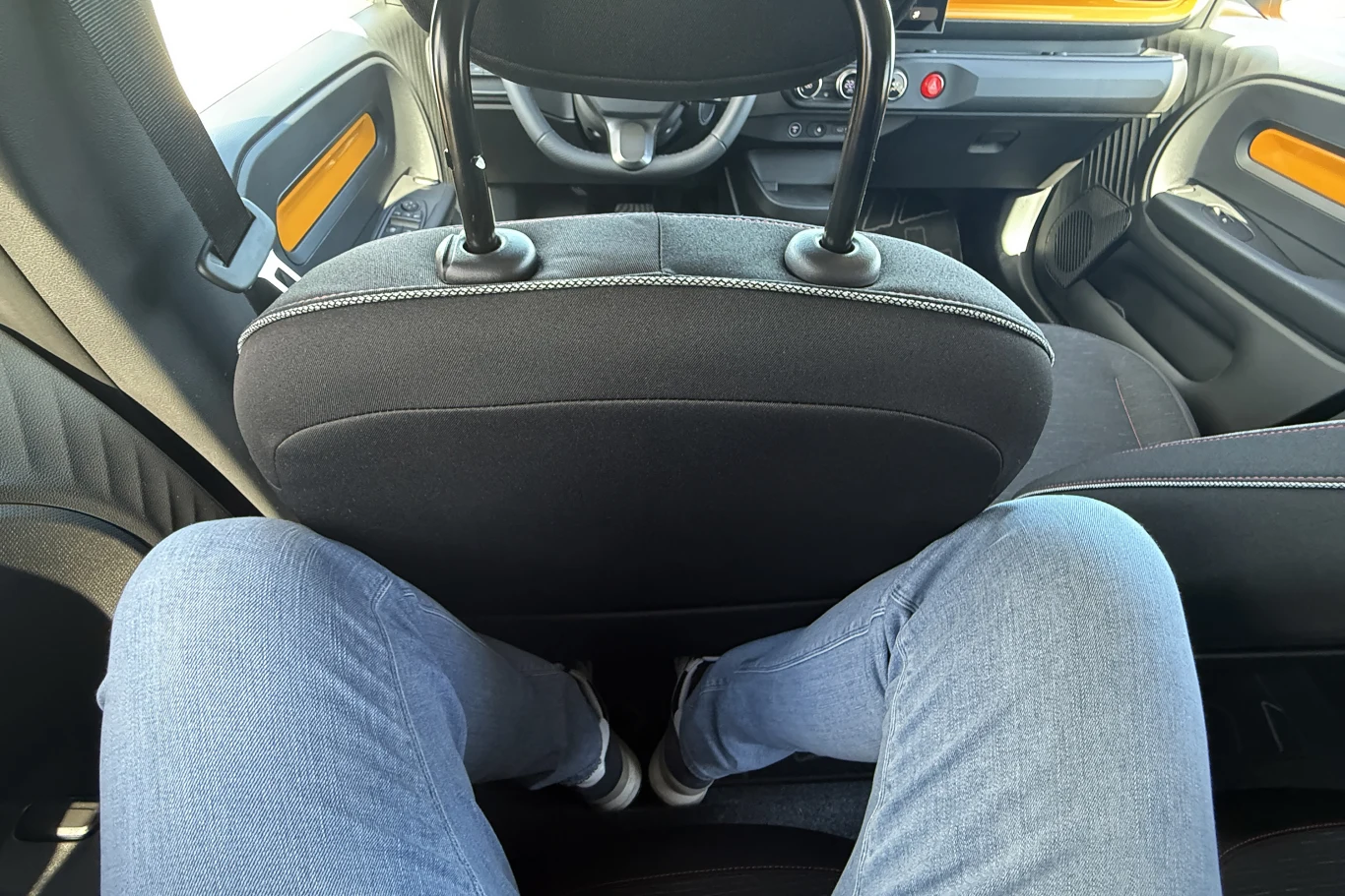 osoba siedząca na tylnej kanapie samochodu z widocznym ograniczonym miejscem na nogi, przedni fotel bardzo blisko kolan, minimalistyczne wnętrze pojazdu z czarno-pomarańczowymi elementami