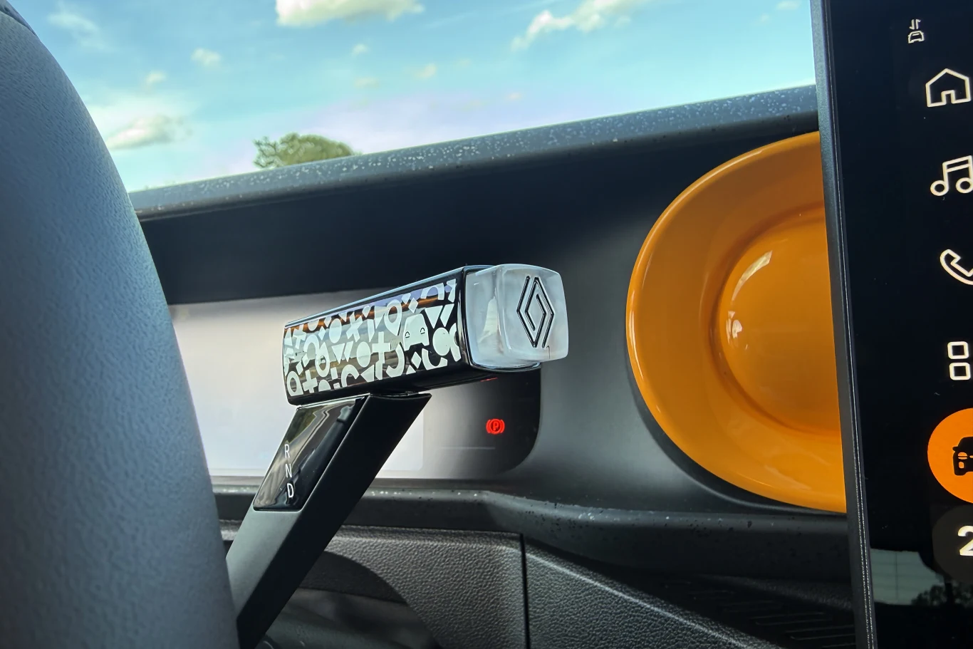 przełącznik zmiany biegów ozdobiony geometrycznym wzorem oraz logo Renault, żółty okrągły element na desce rozdzielczej obok, fragment kierownicy i nowoczesny wyświetlacz dotykowy z ikonami