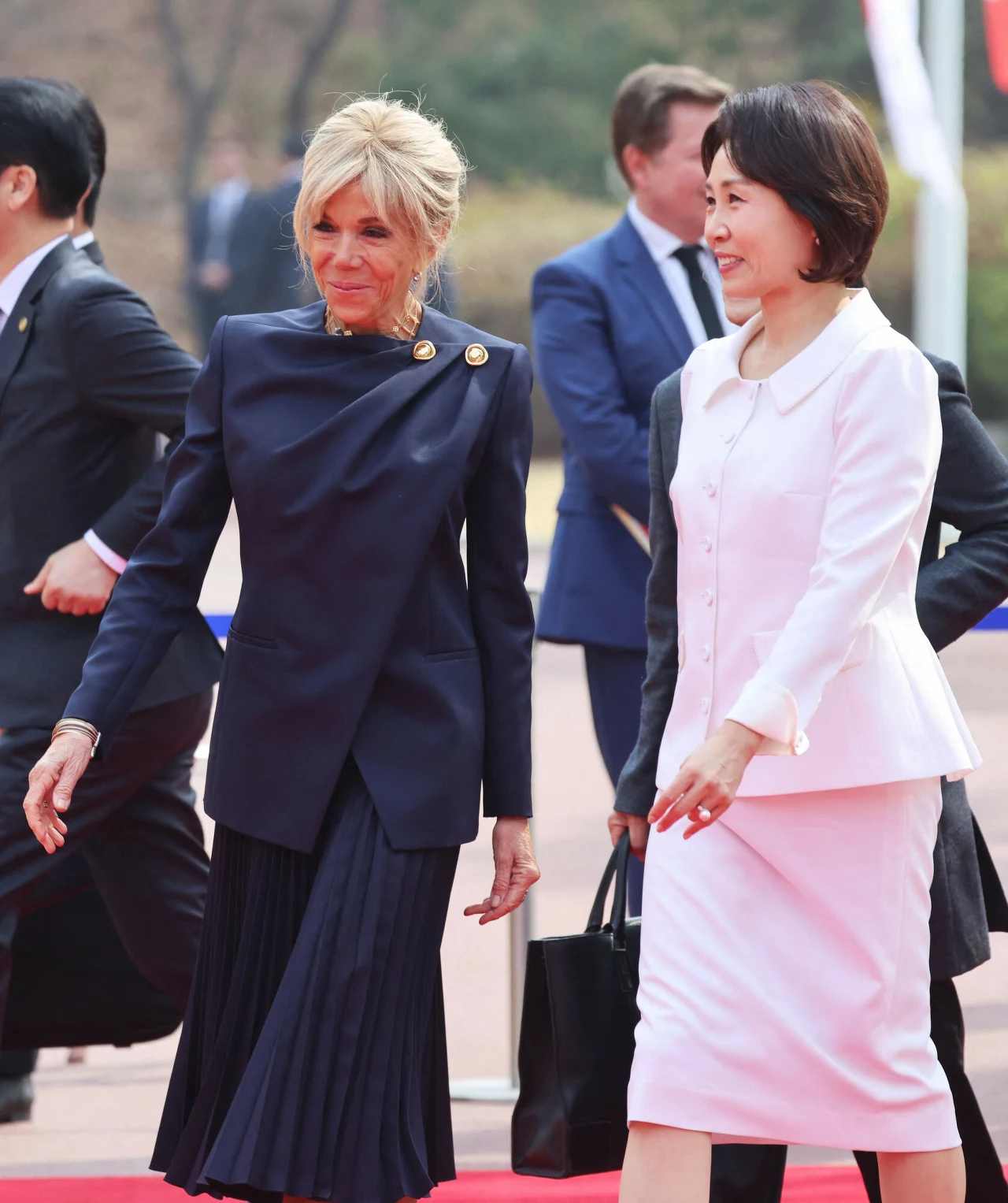 Dwie elegancko ubrane kobiety idą obok siebie po czerwonym dywanie, jedna w granatowym kostiumie, druga w jasnoróżowym komplecie, uśmiechnięte, otoczone przez osoby w garniturach i formalnej scenerii.