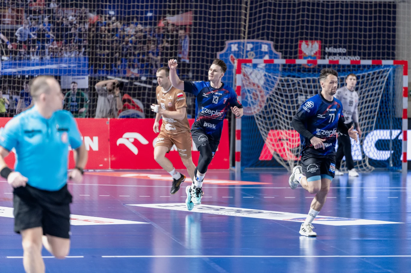 zawodnik w granatowym stroju z uniesioną ręką biegnie po parkiecie hali sportowej, obok niego znajduje się przeciwnik w jasnozłotym stroju oraz drugi zawodnik w niebieskim stroju, na pierwszym planie widoczny jest sędzia z gwizdkiem, w tle bramka i kib...