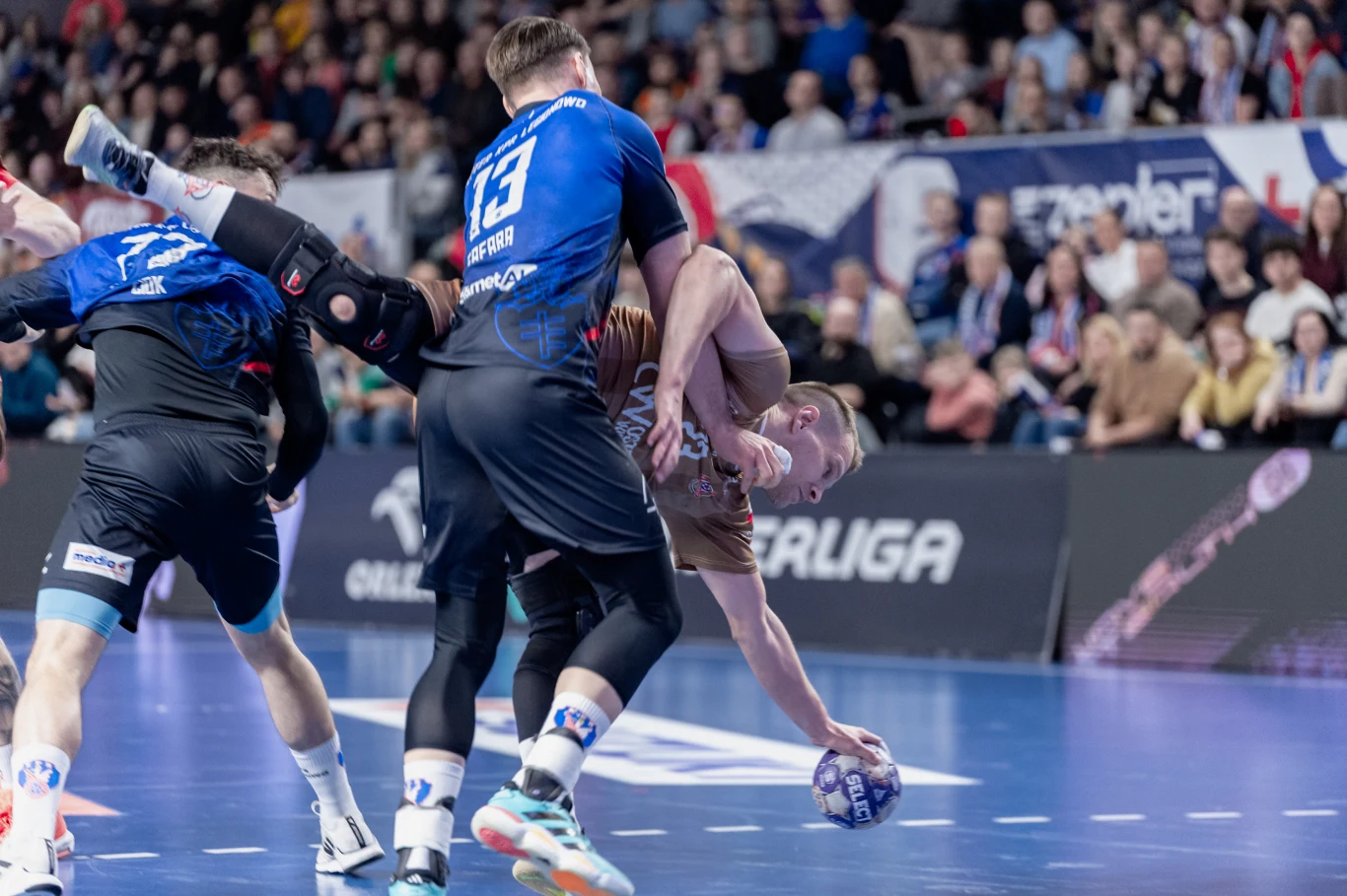 dynamiczna akcja podczas meczu piłki ręcznej, zawodnik w brązowej koszulce walczy o piłkę, będąc zatrzymywanym przez rywala w niebieskim stroju, intensywność wysiłku sportowców podkreśla ich postawa i kontakt fizyczny, w tle widoczna zaangażowana publi...