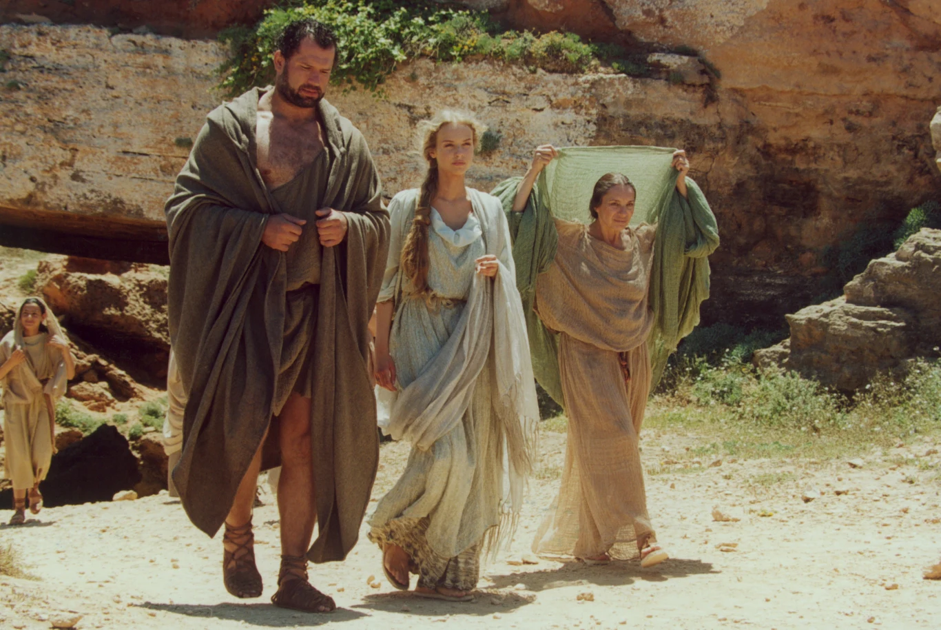 Trzy osoby w antycznych strojach idą razem po piaszczystej ścieżce w skalistym otoczeniu; mężczyzna w długim płaszczu idzie obok dwóch kobiet, z których jedna trzyma przezroczystą tkaninę nad głową dla ochrony przed słońcem.