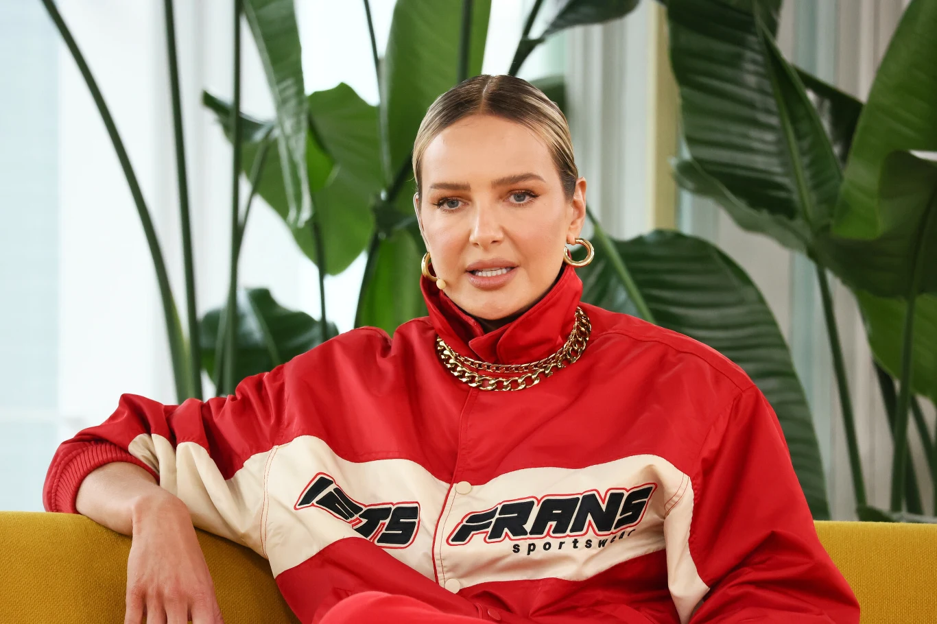 kobieta w czerwonej sportowej kurtce z napisem FRAINS sportswear siedzi na żółtej sofie, ma spięte włosy i nosi złoty łańcuch oraz duże kolczyki, w tle widoczne zielone liście dużych roślin