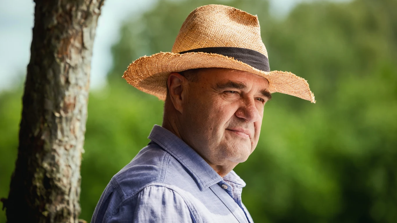 mężczyzna w słomkowym kapeluszu i błękitnej koszuli stoi na tle rozmytego, zielonego krajobrazu, obok pnia drzewa, patrząc w dal