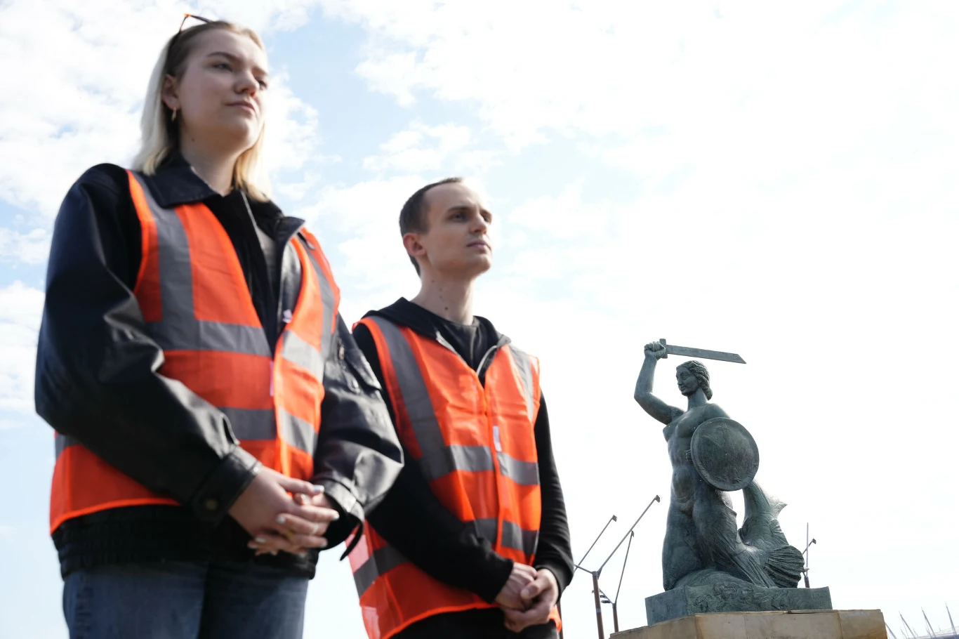 dwie osoby w pomarańczowych kamizelkach odblaskowych stojące obok siebie na tle pomnika Syrenki Warszawskiej