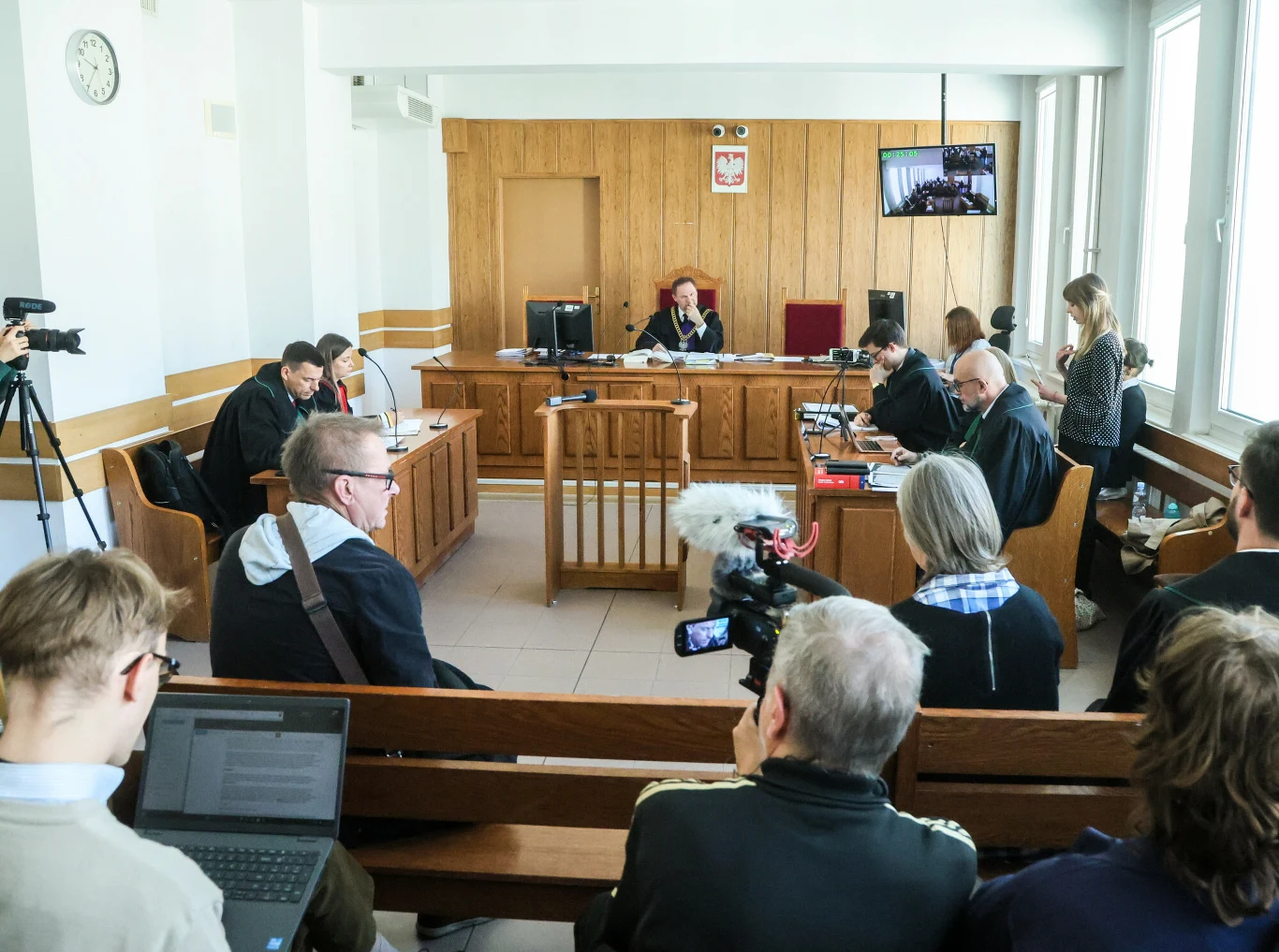 sala sądowa z sędzią siedzącą za stołem sędziowskim, obrońcami i oskarżycielem po bokach, publiczność i dziennikarze z kamerami oraz sprzętem do nagrywania siedzący na ławach naprzeciwko sędziego