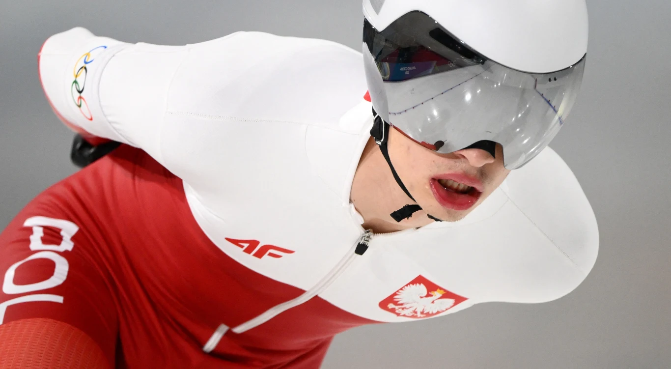 polski panczenista w biało-czerwonym stroju z godłem Polski i napisem 'POL', pochylony do przodu podczas jazdy, w kasku i okularach ochronnych