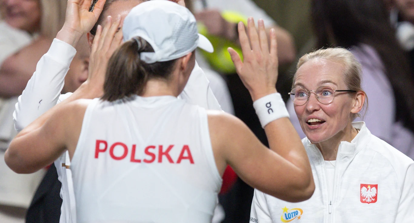 Tenisistka w stroju z napisem 'Polska' i czapce przybija piątkę z uśmiechniętą kobietą w białej kurtce z polskim godłem i logo Lotto, obie osoby wyrażają entuzjazm i radość po sportowym wydarzeniu.
