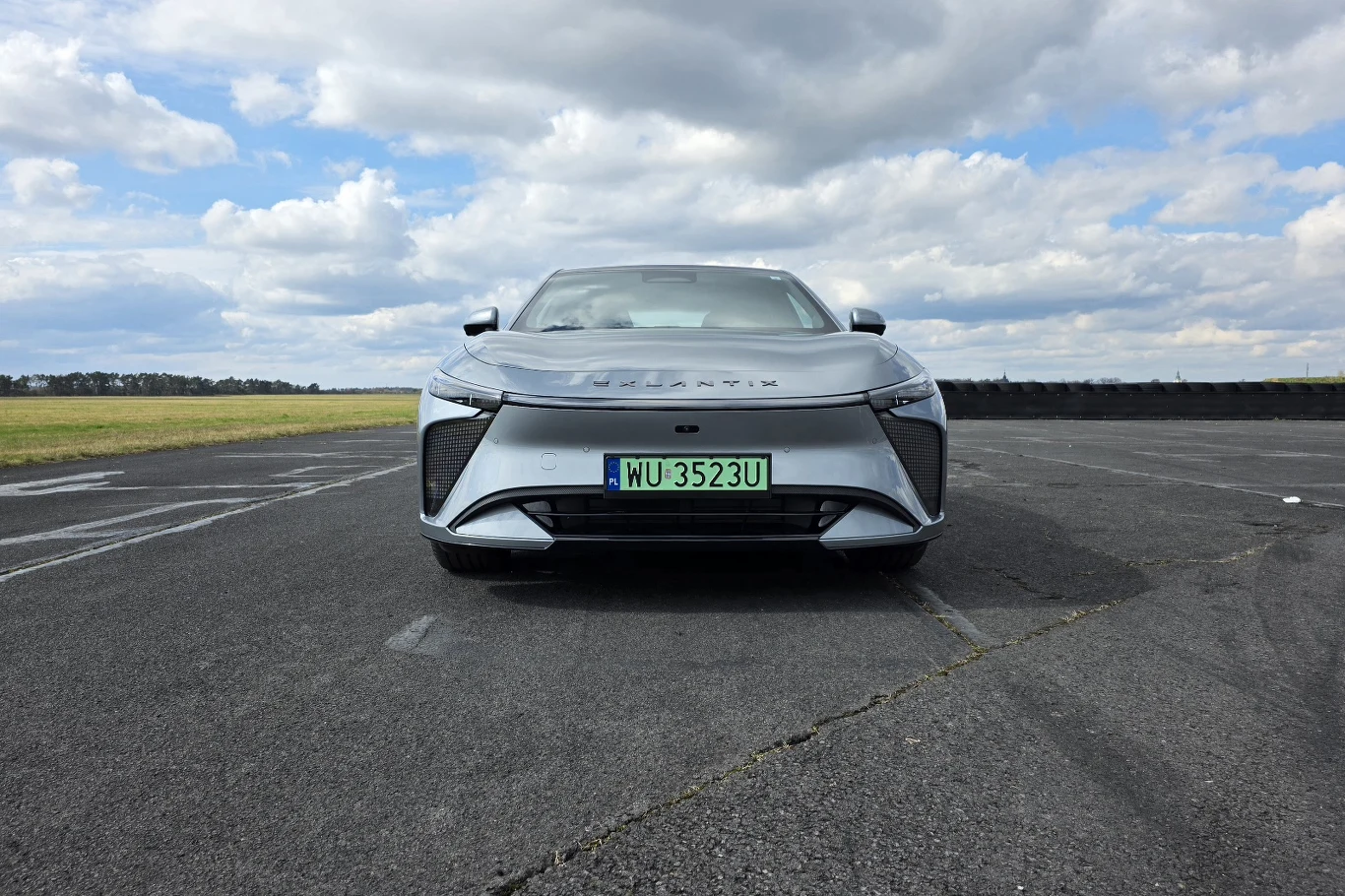 srebrny samochód elektryczny marki Lexus ustawiony na pustym asfalcie z widocznymi tablicami rejestracyjnymi, w tle rozpościera się otwarta przestrzeń i niebo z licznymi chmurami