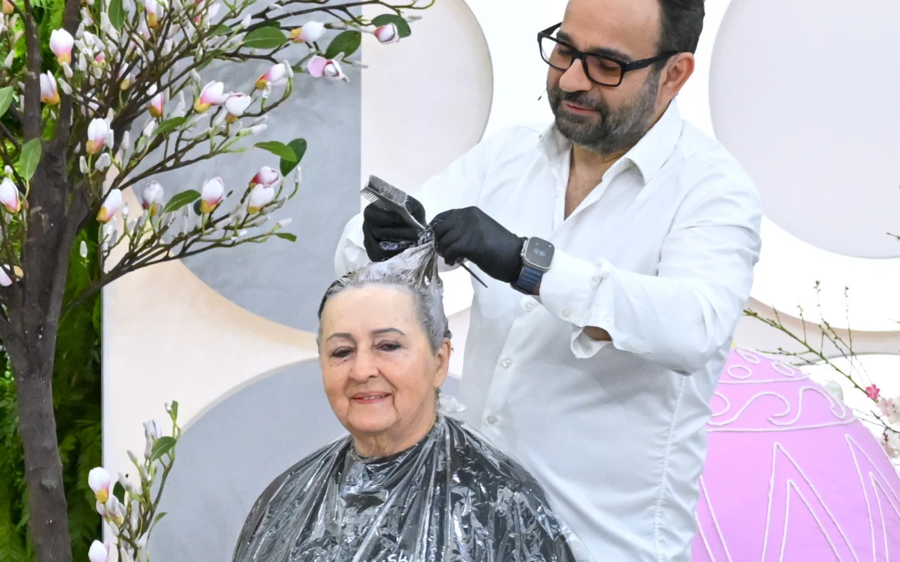 starsza kobieta siedzi z uśmiechem na twarzy, fryzjer w okularach nakłada jej farbę na włosy, wokół widać dekoracje z kwitnącymi gałązkami i pastelowymi elementami