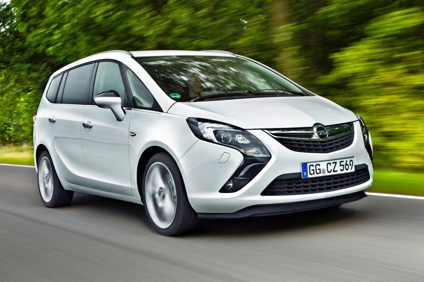 biały Opel Zafira poruszający się po asfaltowej drodze, otoczony gęstą zielenią drzew w tle