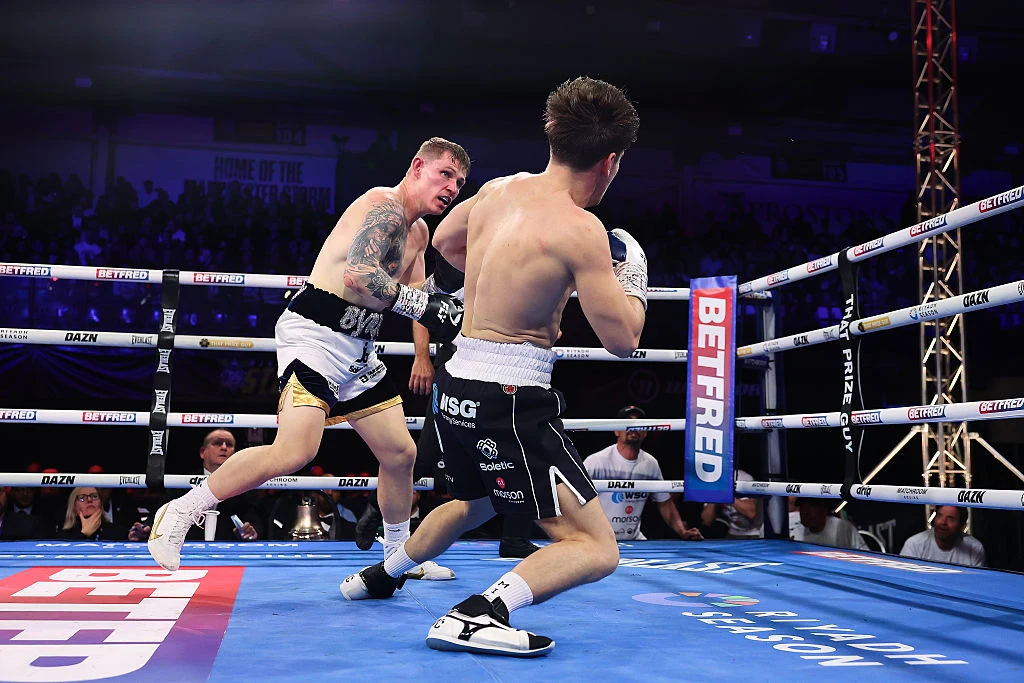 dwóch bokserów w ringu, jeden z nich pada na matę po silnym ciosie przeciwnika, wokół ring otoczony linami, a w tle widoczni sędziowie i publiczność