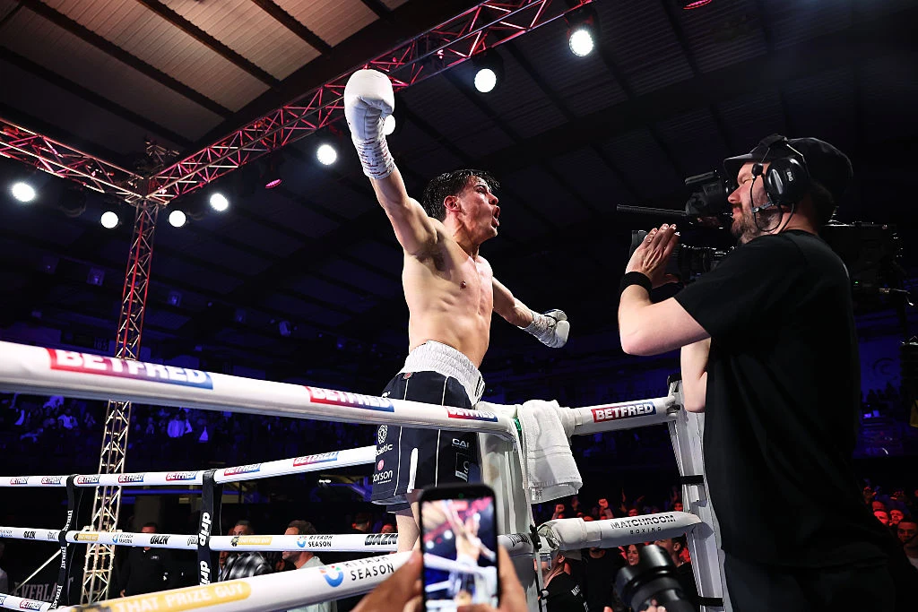 Bokser w uniesionych rękawicach stoi na linach ringu, świętując zwycięstwo, podczas gdy kamerzysta rejestruje jego reakcję, a w tle widoczna jest publiczność i oświetlenie hali.