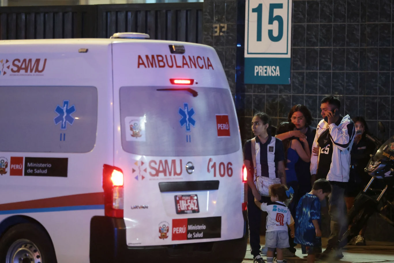 ambulans należący do peruwiańskiego Ministerstwa Zdrowia zaparkowany obok grupy ludzi, wśród których znajduje się kobieta trzymająca dziecko za rękę, wszyscy stoją w pobliżu wejścia oznaczonego numerem 15