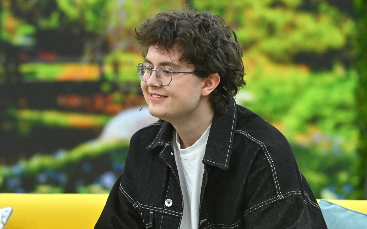 Młody mężczyzna z kręconymi włosami i okularami, ubrany w czarną kurtkę z białym t-shirtem, siedzi na żółtej sofie i uśmiecha się, w tle rozmyte zielone rośliny.