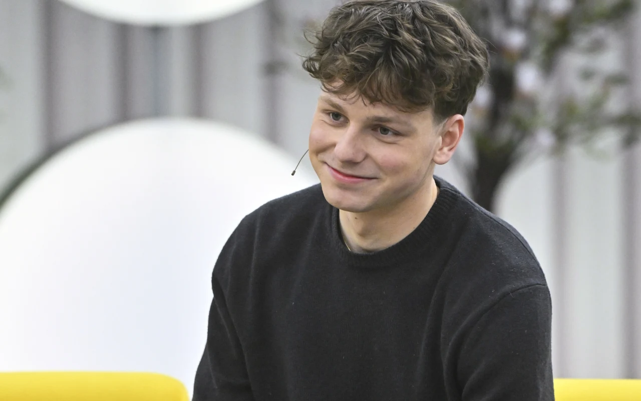 Wojciech Czuba w "halo tu polsat" Młody mężczyzna z mikrofonem przypiętym do ucha siedzi na żółtej sofie, ubrany w ciemny sweter, uśmiecha się lekko, w tle rozmyte okrągłe światła i rośliny.