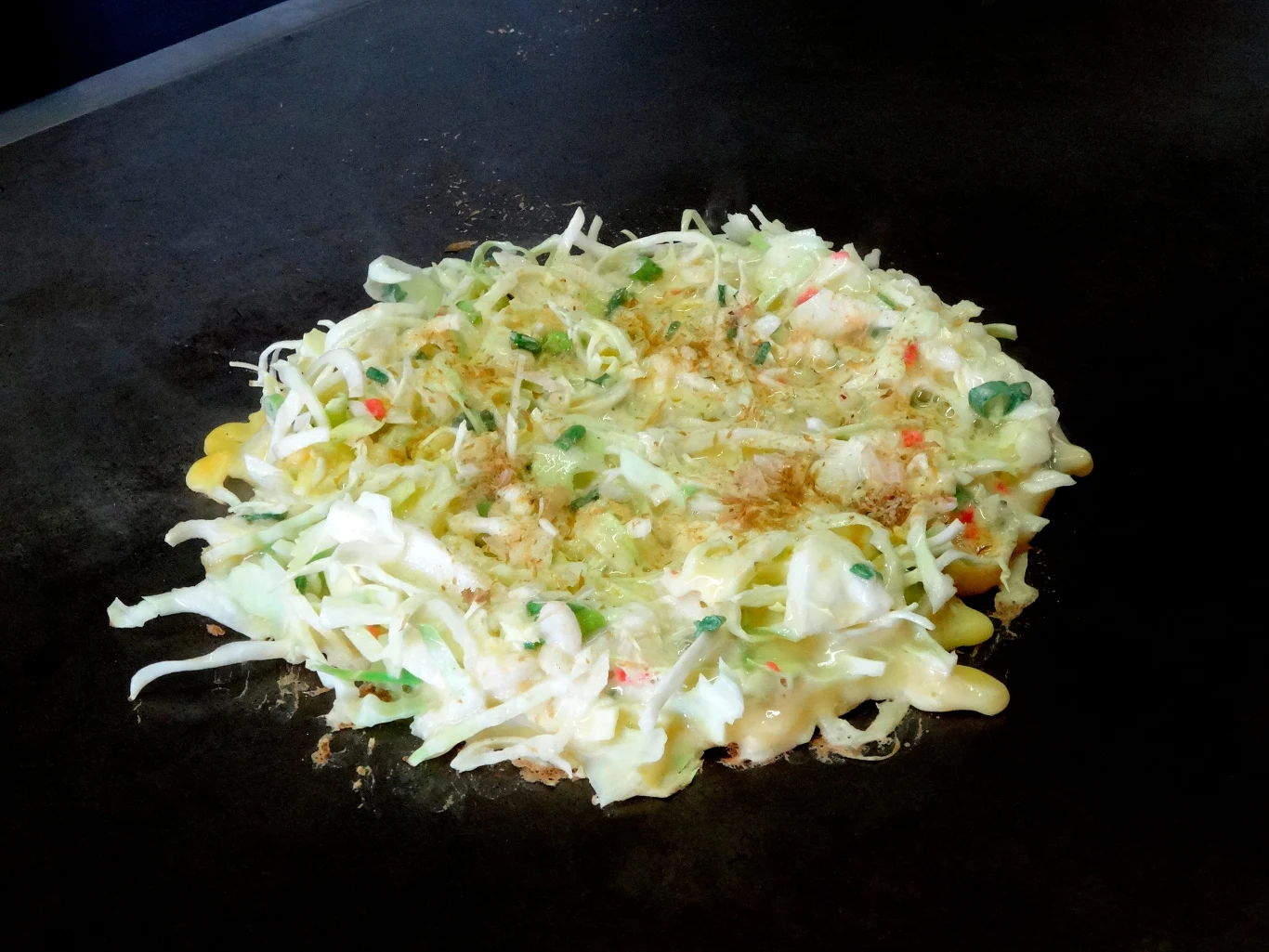 Japoński placek okonomiyaki z warstwą świeżej kapusty, posiekanymi szczypiorkami oraz kawałkami czerwonej papryki, smażony na płaskiej żeliwnej płycie.