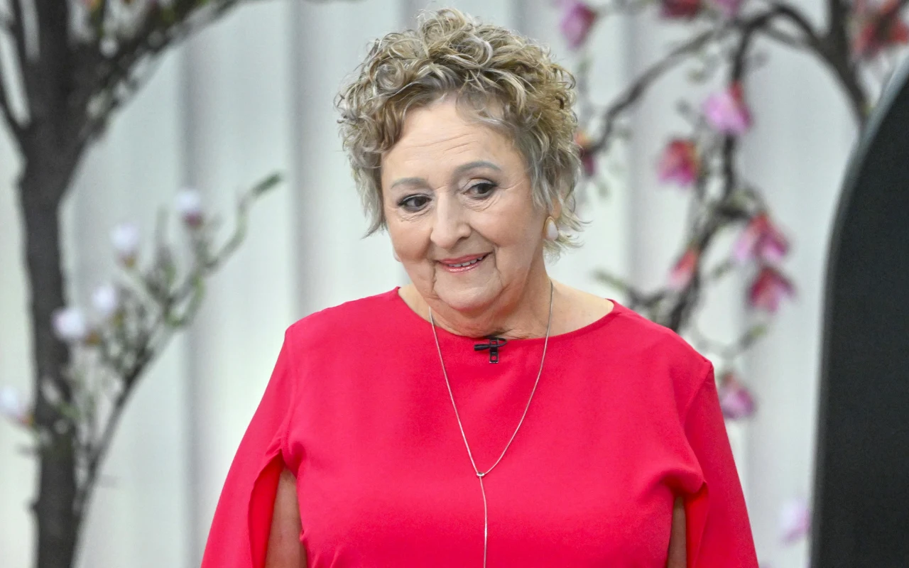 starsza kobieta z krótkimi, kręconymi włosami w czerwonej sukience stoi na tle ozdobnych gałęzi z różowymi kwiatami