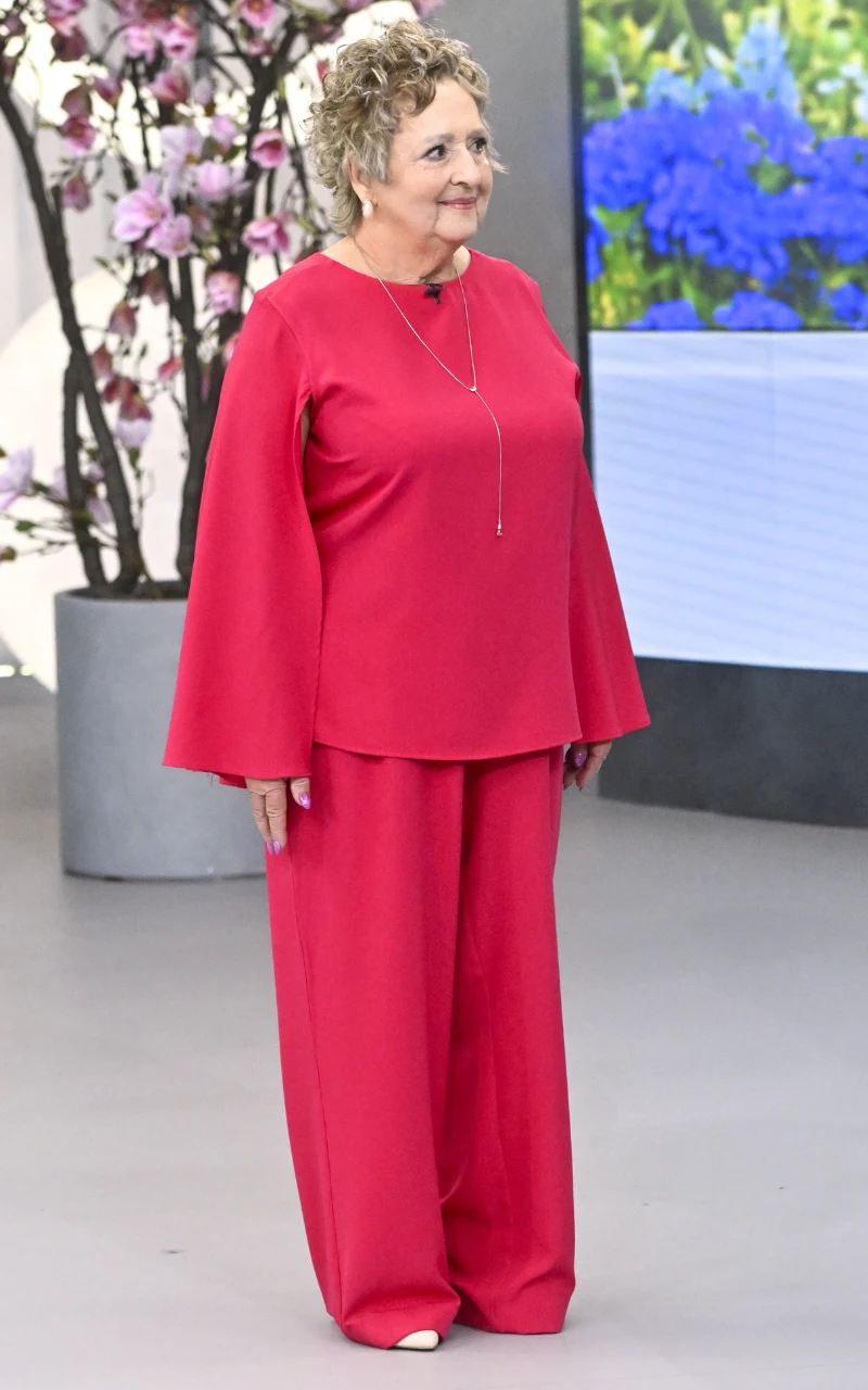 starsza kobieta z krótkimi kręconymi włosami ubrana w luźny, czerwony komplet z szerokimi spodniami i długą bluzką z rozkloszowanymi rękawami, nosząca długi naszyjnik, stojąca na tle dekoracji z kwiatami i ekranu z wyświetlonymi niebieskimi roślinami