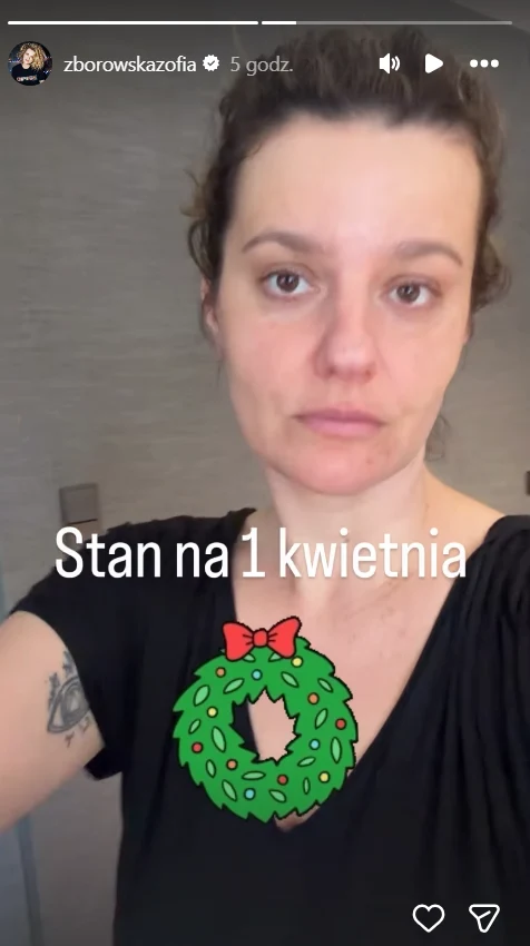 kobieta o poważnym wyrazie twarzy z ciemnymi włosami spiętymi do góry, ubrana w czarną bluzkę z widocznym tatuażem na lewym ramieniu, na środku napisy „Stan na 1 kwietnia” oraz grafika świątecznego wieńca z czerwoną kokardką