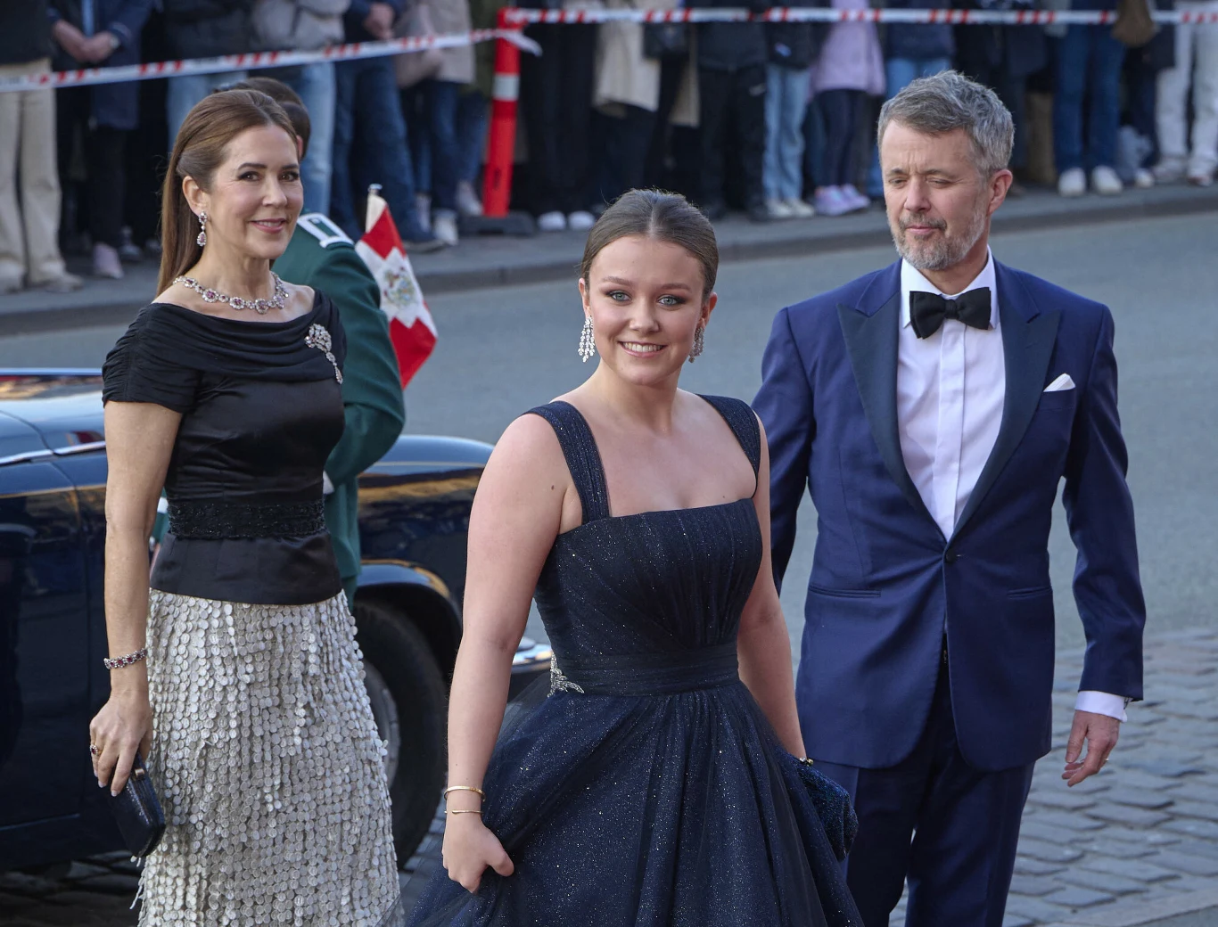 Trzy elegancko ubrane osoby wchodzące po bruku – kobieta w ciemnej sukni z bogatą spódnicą, młoda kobieta w granatowej sukni balowej oraz mężczyzna w granatowym garniturze z muchą; w tle samochód i tłum obserwujących ludzi za barierkami.