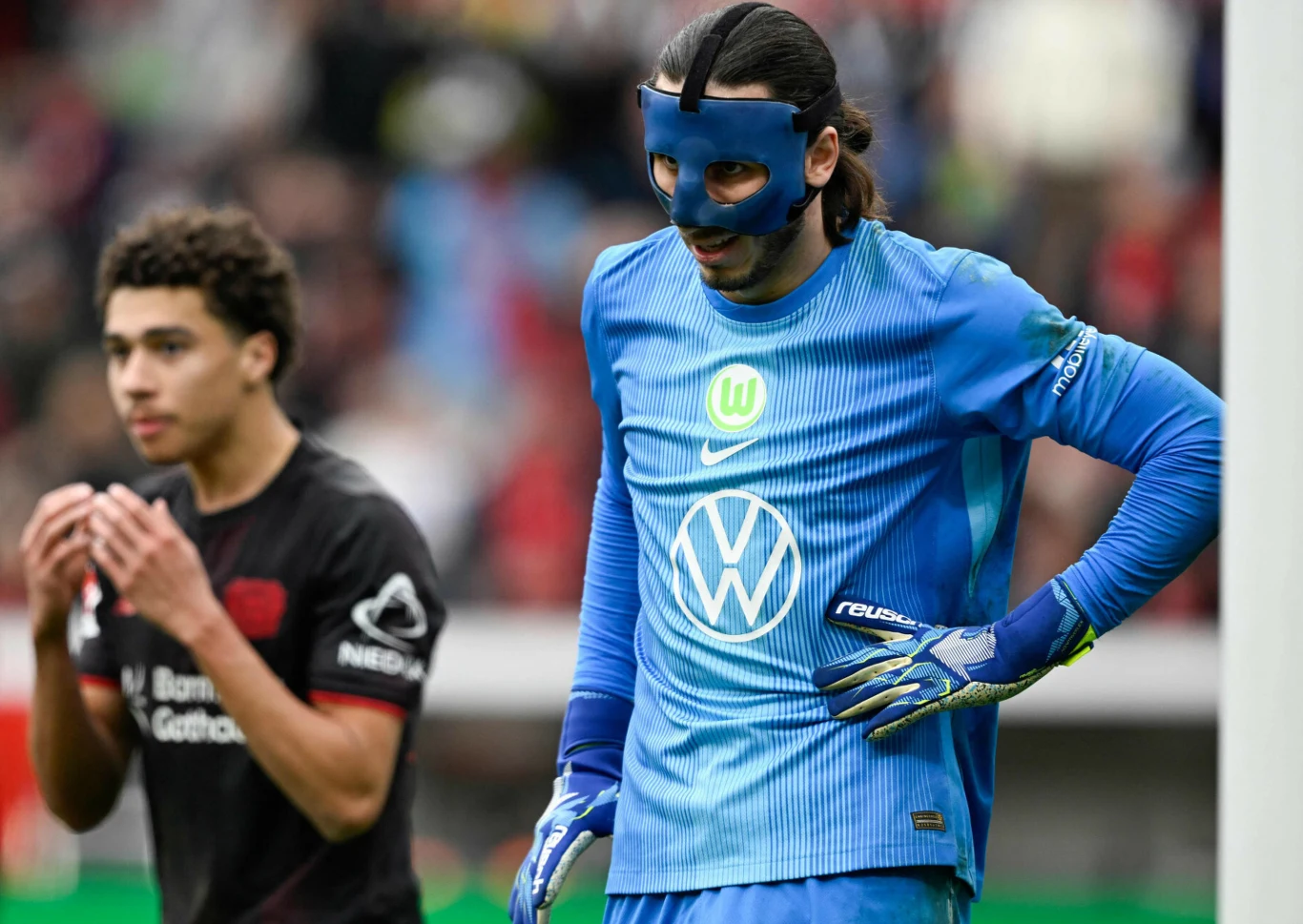 bramkarz w niebieskim stroju i czarnej masce ochronnej na twarzy stoi przy słupku bramki, obok zawodnik drużyny przeciwnej w czarno-czerwonym stroju z widocznym logo sponsora na rękawie