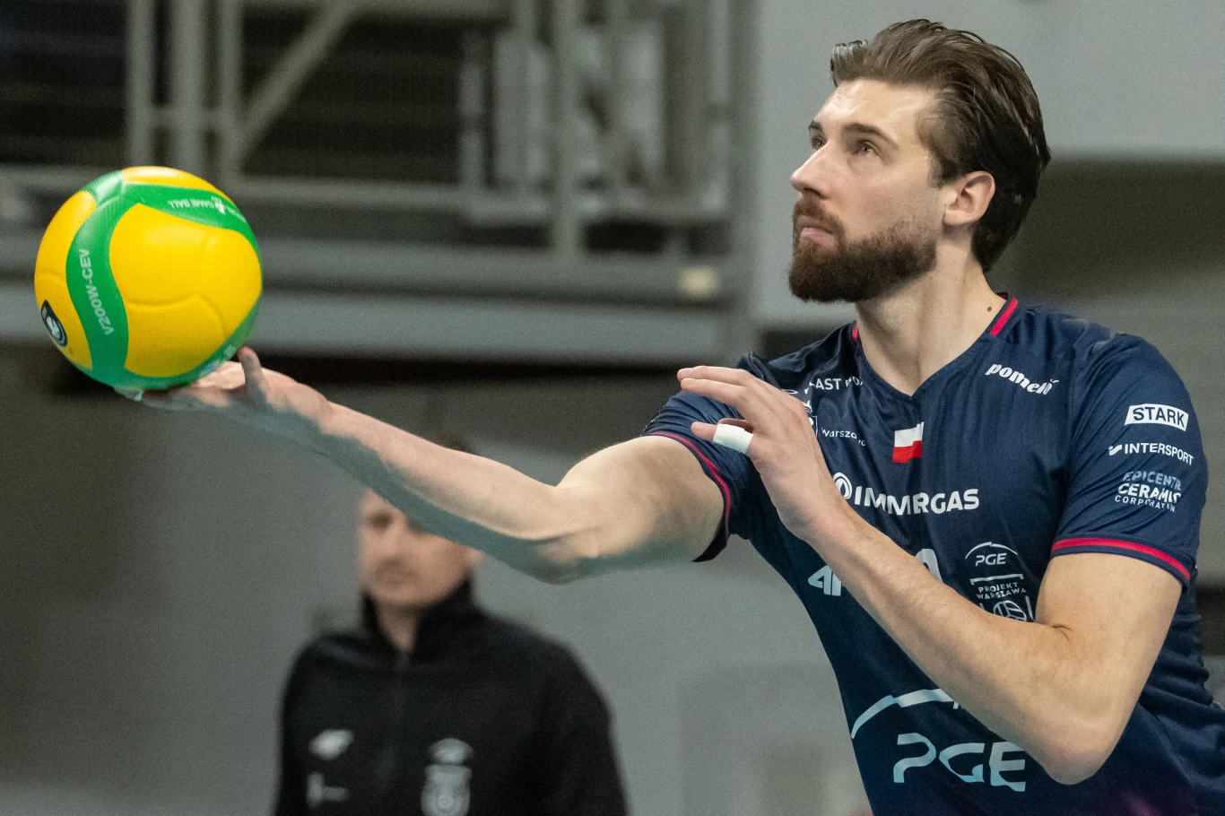 siatkarz w granatowym stroju serwuje piłkę podczas meczu, skupiony wyraz twarzy i dynamiczna postawa podkreślają moment koncentracji i gotowości do gry