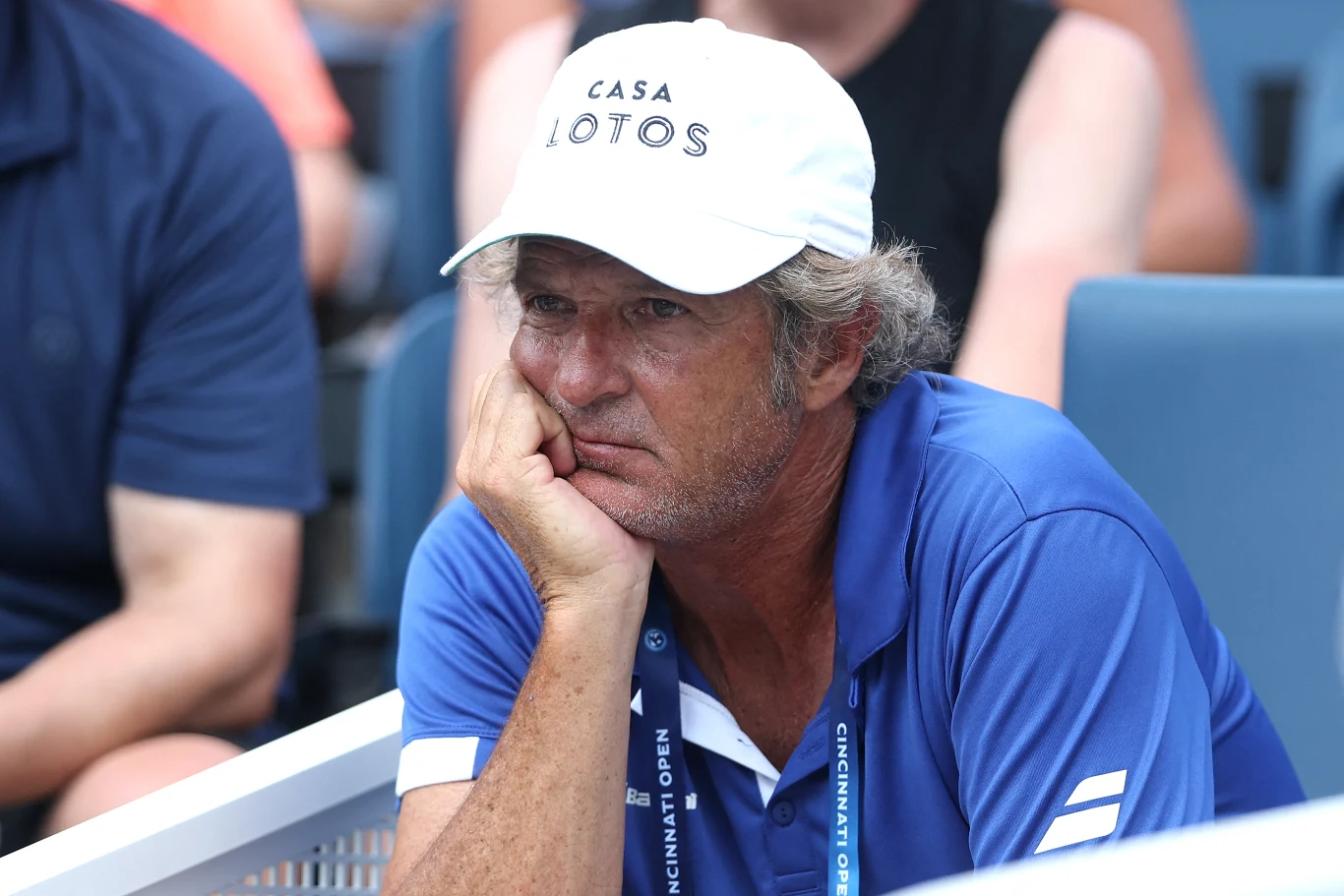 Mężczyzna w czapce z daszkiem z napisem 'CASA LOTOS', ubrany w niebieską koszulkę polo z białymi detalami, siedzi oparty o rękę z zamyśloną miną, w tle widoczne są rozmyte sylwetki innych osób.