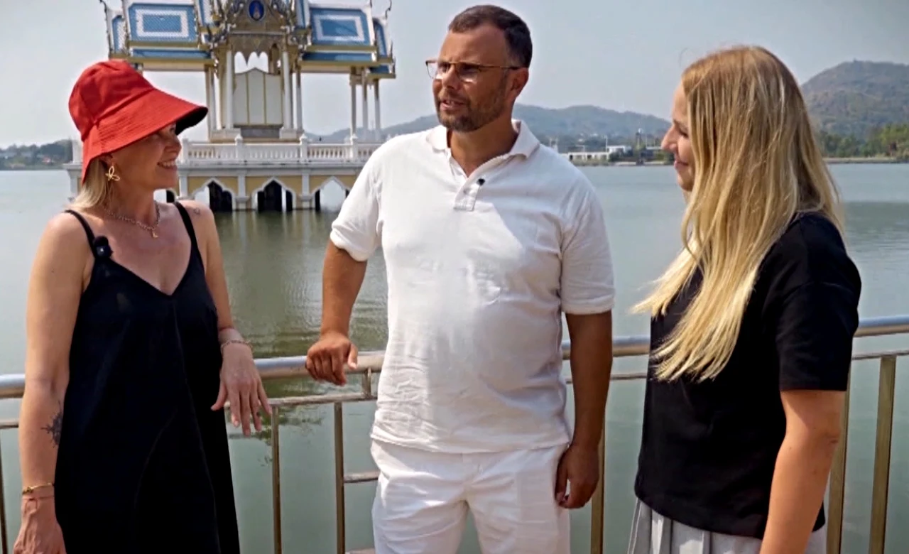 Święta Polaków za granicą Trzy osoby rozmawiają przed malowniczym pawilonem nad wodą; kobieta w czerwonym kapeluszu, mężczyzna w bieli i kobieta.