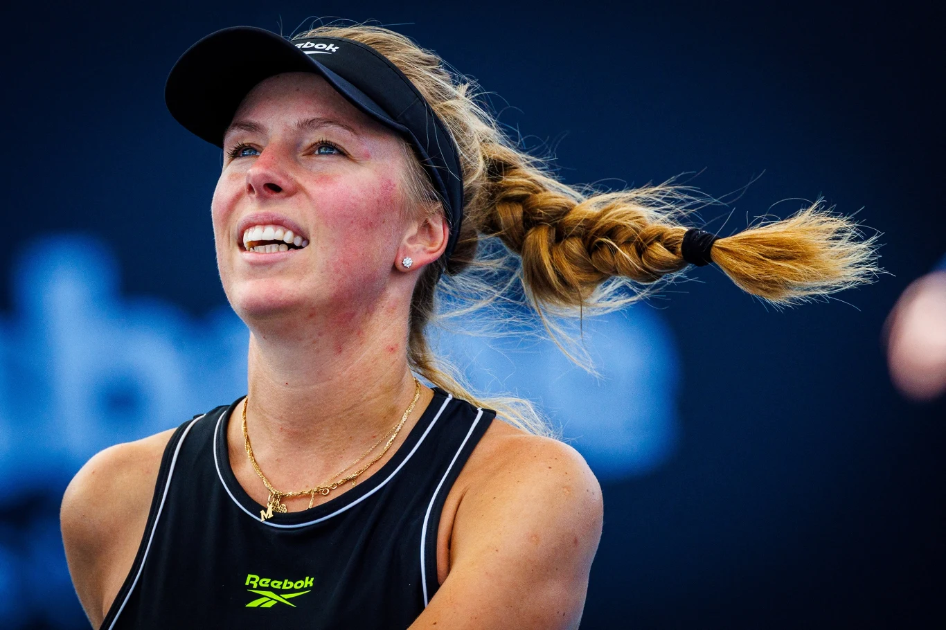 Magdalena Fręch Kobieta w sportowym stroju z widocznym logiem Reebok, uśmiechnięta, z opaską na głowie i długim, splecionym warkoczem dynamicznie odchylonym w bok, tło rozmyte w niebieskich odcieniach.