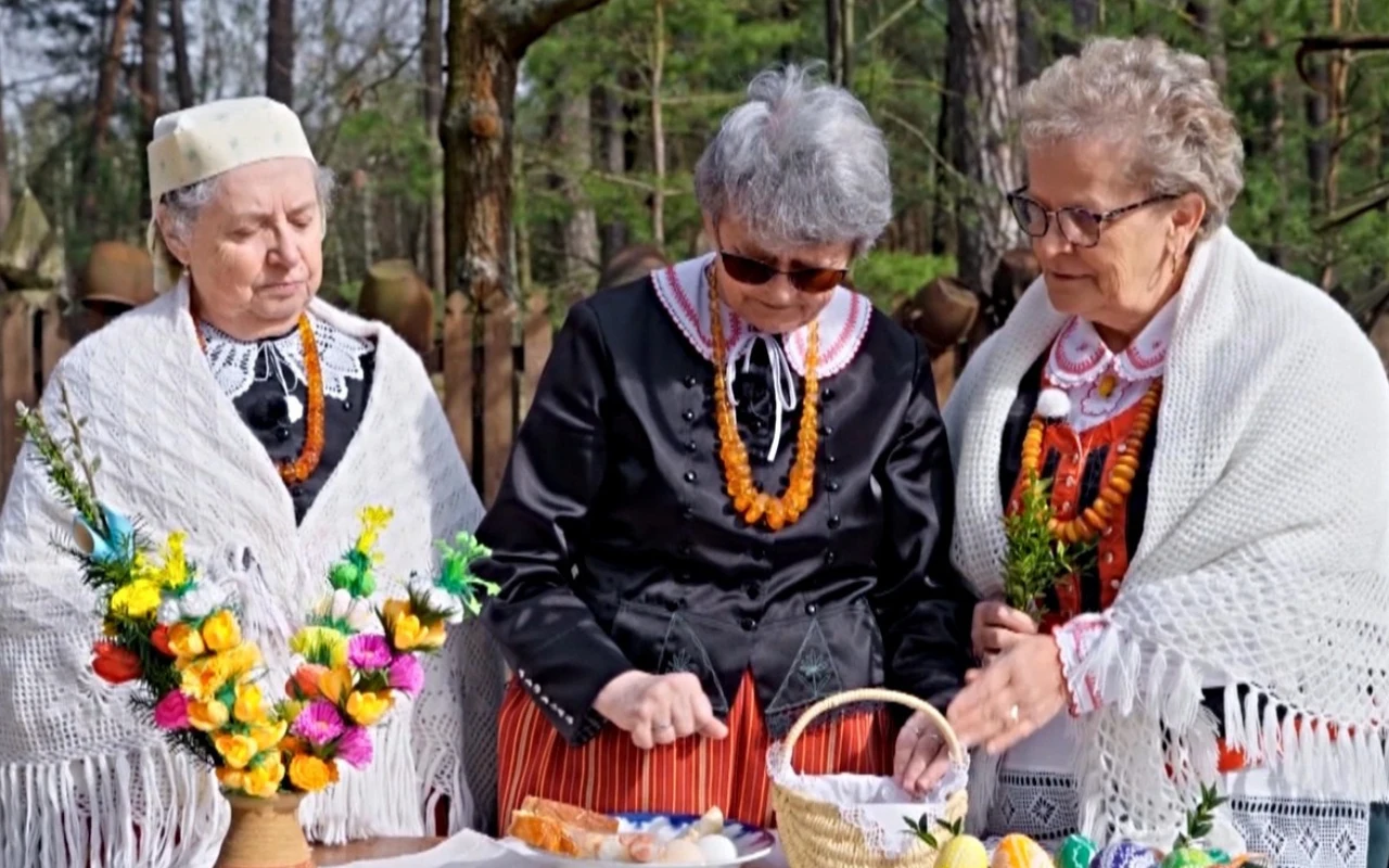 Wielkanoc na Kurpiach Trzy starsze kobiety w kurpiowskich strojach ludowych przy stole z palmami, pisankami i koszykiem święconki.