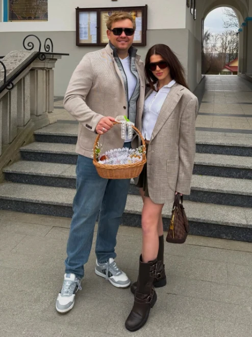 Para młodych osób w modnych ubraniach stoi razem na schodach przed budynkiem, mężczyzna trzyma w ręce ozdobiony koszyk z dekoracjami wielkanocnymi, oboje mają na sobie okulary przeciwsłoneczne.