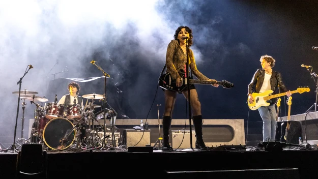 Trójka muzyków występuje na scenie oświetlonej jasnym światłem, centralnie znajduje się wokalistka grająca na gitarze elektrycznej, po lewej perkusista, po prawej basista; otacza ich dym sceniczny i ciemne tło, co podkreśla dynamiczny charakter koncertu.