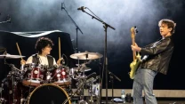 Perkusista w ciemnych włosach gra dynamicznie na dużym, rozbudowanym zestawie perkusyjnym, a obok niego gitarzysta w skórzanej kurtce, pochylony do przodu, energicznie gra na gitarze elektrycznej; w tle unształcona scenografia i wyraźny mikrofon scenic...
