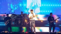 zespół muzyczny występujący na scenie, wokalista śpiewający do mikrofonu, gitarzysta, basista oraz perkusista grający na żywo, dynamiczne oświetlenie LED i kolorowe wizualizacje w tle