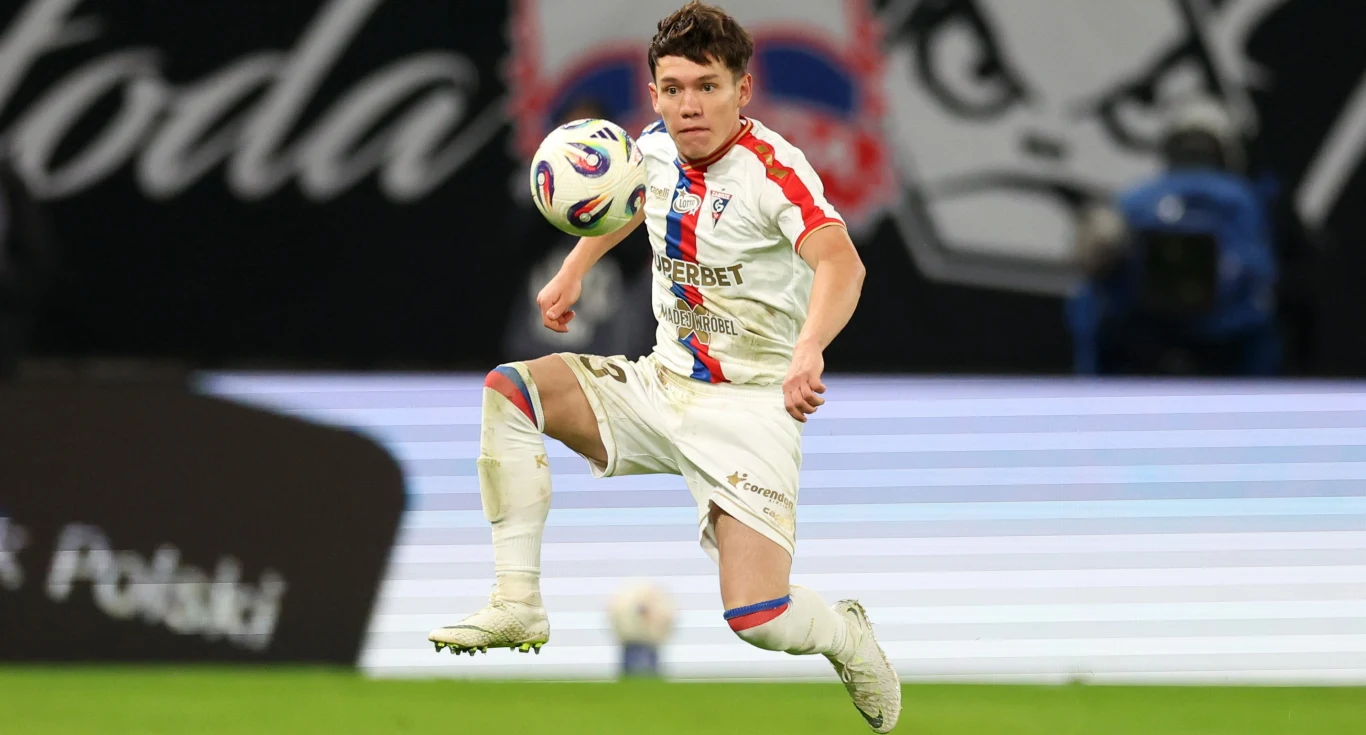 piłkarz w białym stroju sportowym unosi się w powietrzu podczas dynamicznego zagrania piłki, skupienie na twarzy i intensywna akcja na tle rozmytego stadionu oraz charakterystycznej grafiki na trybunach