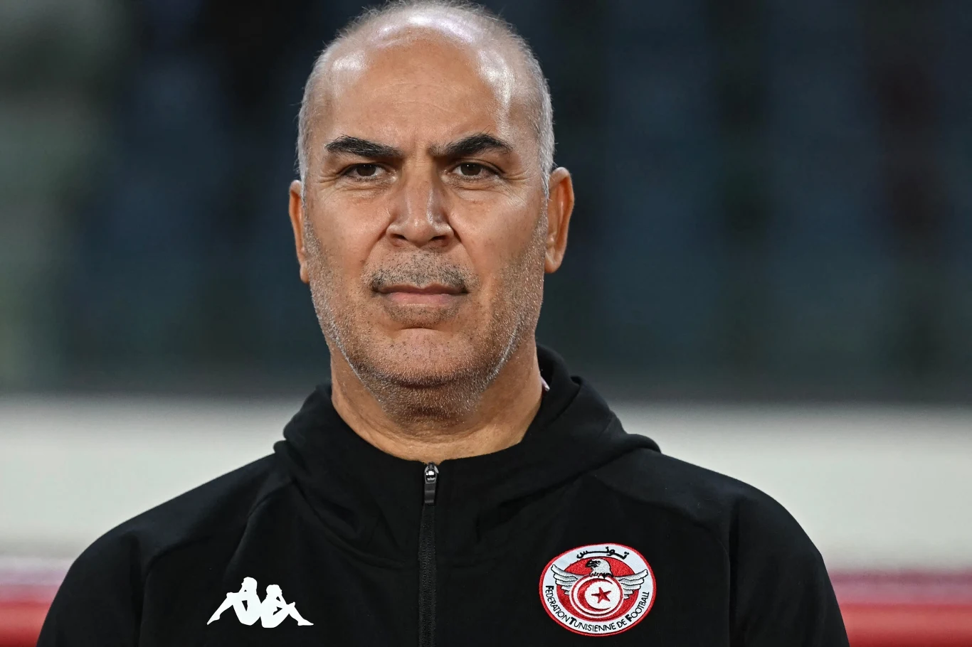 mężczyzna o łysiejących włosach i lekko siwiejącej brodzie, ubrany w czarną bluzę sportową z logo Kappa i herbem tunezyjskiej federacji piłkarskiej, patrzy poważnie prosto przed siebie