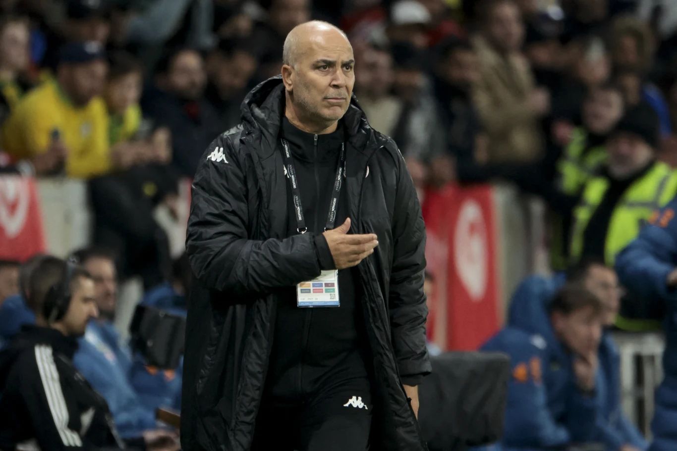 łysy mężczyzna w czarnej kurtce sportowej z identyfikatorem na szyi stoi na tle kibiców na stadionie, skoncentrowany i skupiony