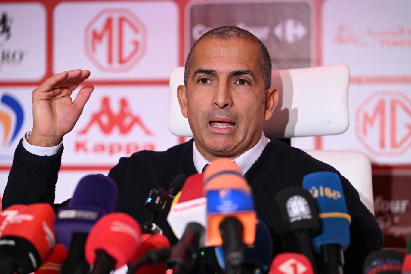 Mężczyzna siedzi na białym krześle, przemawia do mediów, otoczony licznymi mikrofonami różnych stacji telewizyjnych i radiowych.