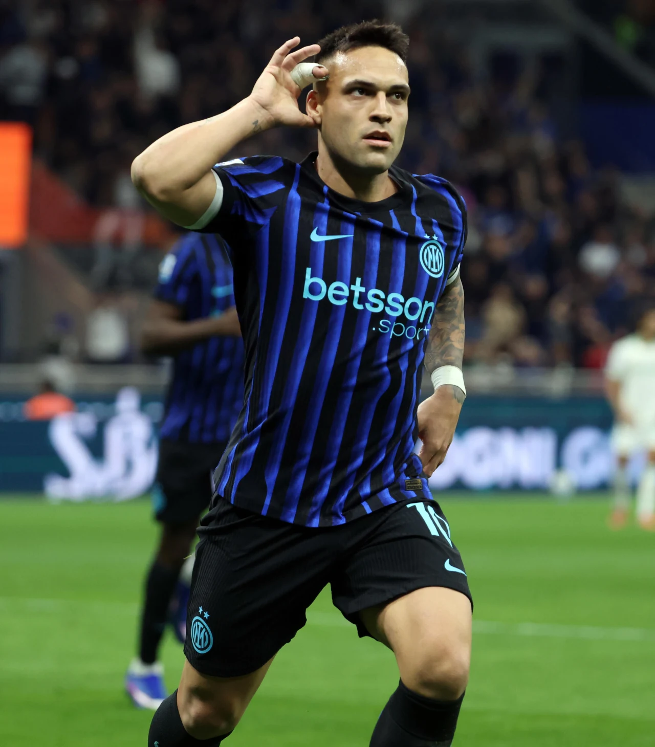 piłkarz w niebiesko-czarnej koszulce Interu Mediolan świętuje strzelenie gola, przykładając dłoń do ucha i biegnąc po murawie na tle rozmytego stadionu