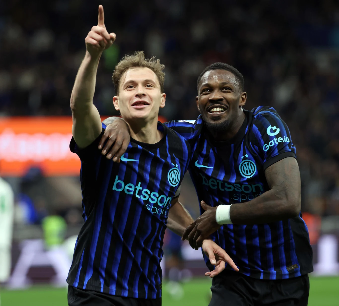 Dwóch piłkarzy Interu Mediolan w niebiesko-czarnych koszulkach świętuje zdobycie gola, obejmując się; jeden z nich z szerokim uśmiechem unosi rękę i wskazuje palcem w górę.