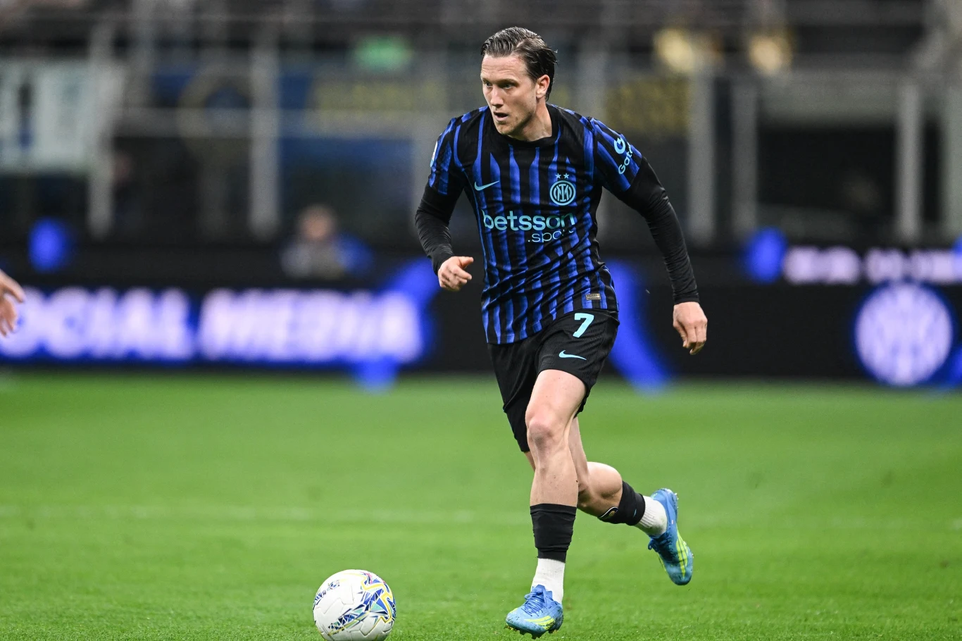 Piłkarz w niebiesko-czarnej koszulce z numerem 7 prowadzi piłkę po zielonej murawie podczas meczu, skupiwszy wzrok na kierunku podania.