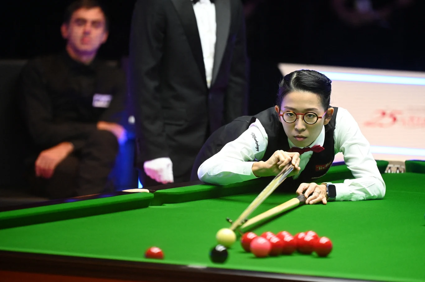 zawodnik w okularach skupiony na wykonaniu uderzenia podczas meczu snookera, precyzyjnie celując kijem bilardowym w białą bilę, na stole widoczne czerwone bile ułożone w pobliżu narożnika, w tle sędzia i obserwujący rywal