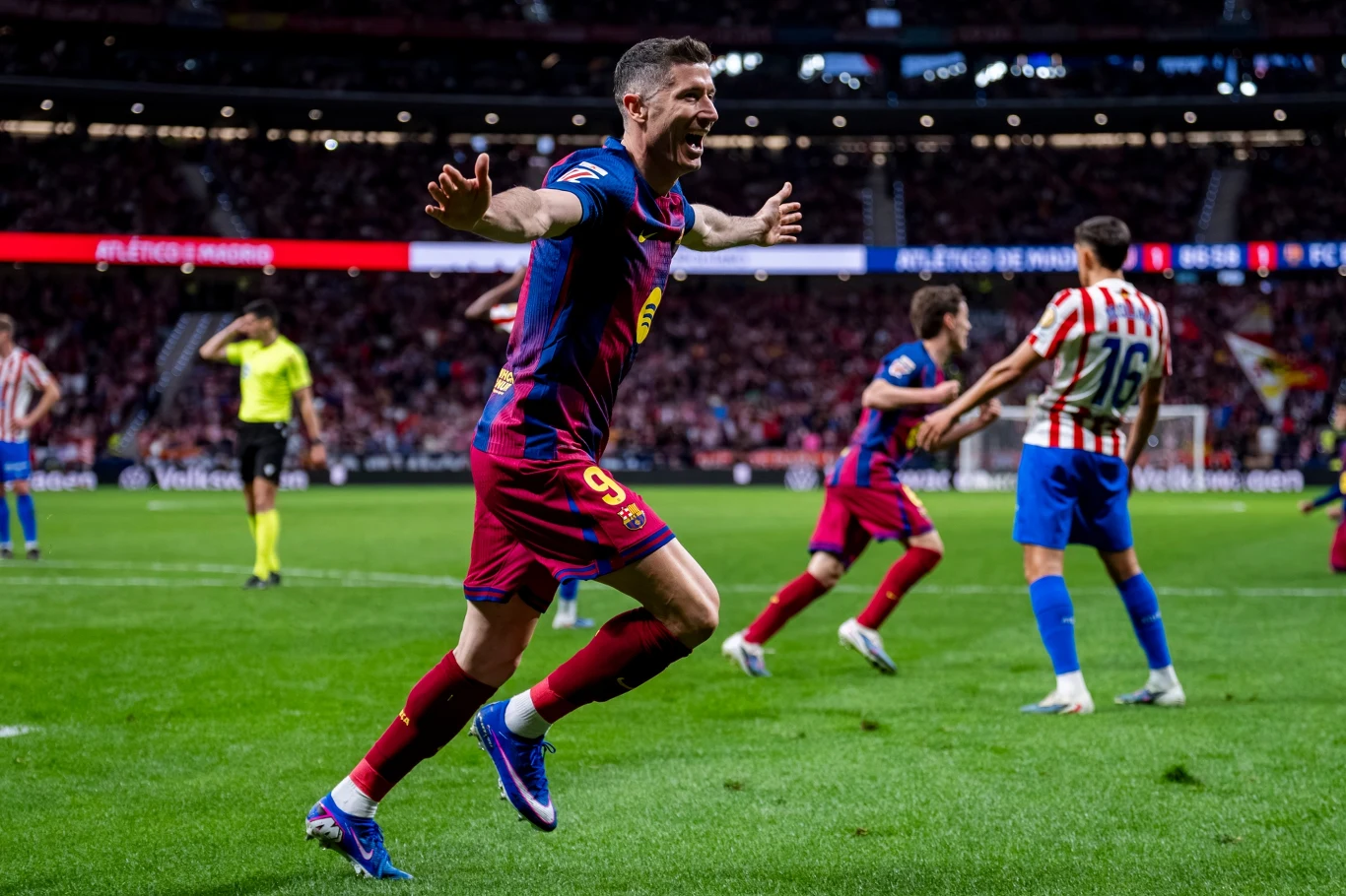 piłkarz w bordowo-granatowym stroju z numerem 9 świętuje zdobycie gola, szeroko rozkładając ręce, podczas gdy na tle stadionu widać zawodników drużyny przeciwnej oraz sędziego