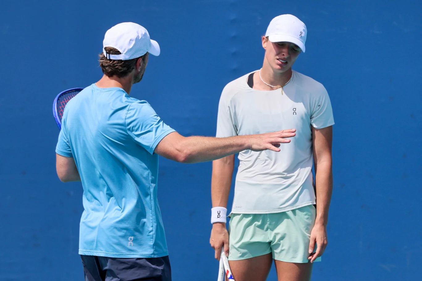 Dwoje tenisistów w jasnych strojach podczas rozmowy na korcie, jedna z osób gestykuluje ręką, wyraźnie tłumacząc coś drugiej osobie, która trzyma rakietę i wygląda na zamyśloną.
