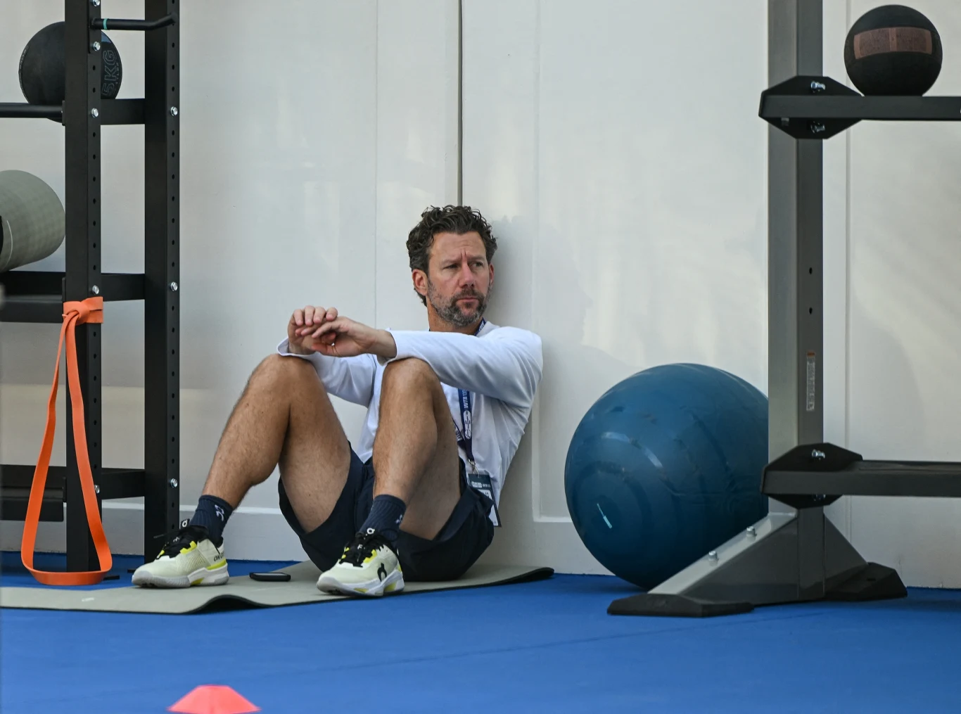 Mężczyzna w sportowym stroju siedzi oparty o ścianę na macie w siłowni, obok niego znajduje się niebieska piłka gimnastyczna oraz stojak na sprzęt sportowy.