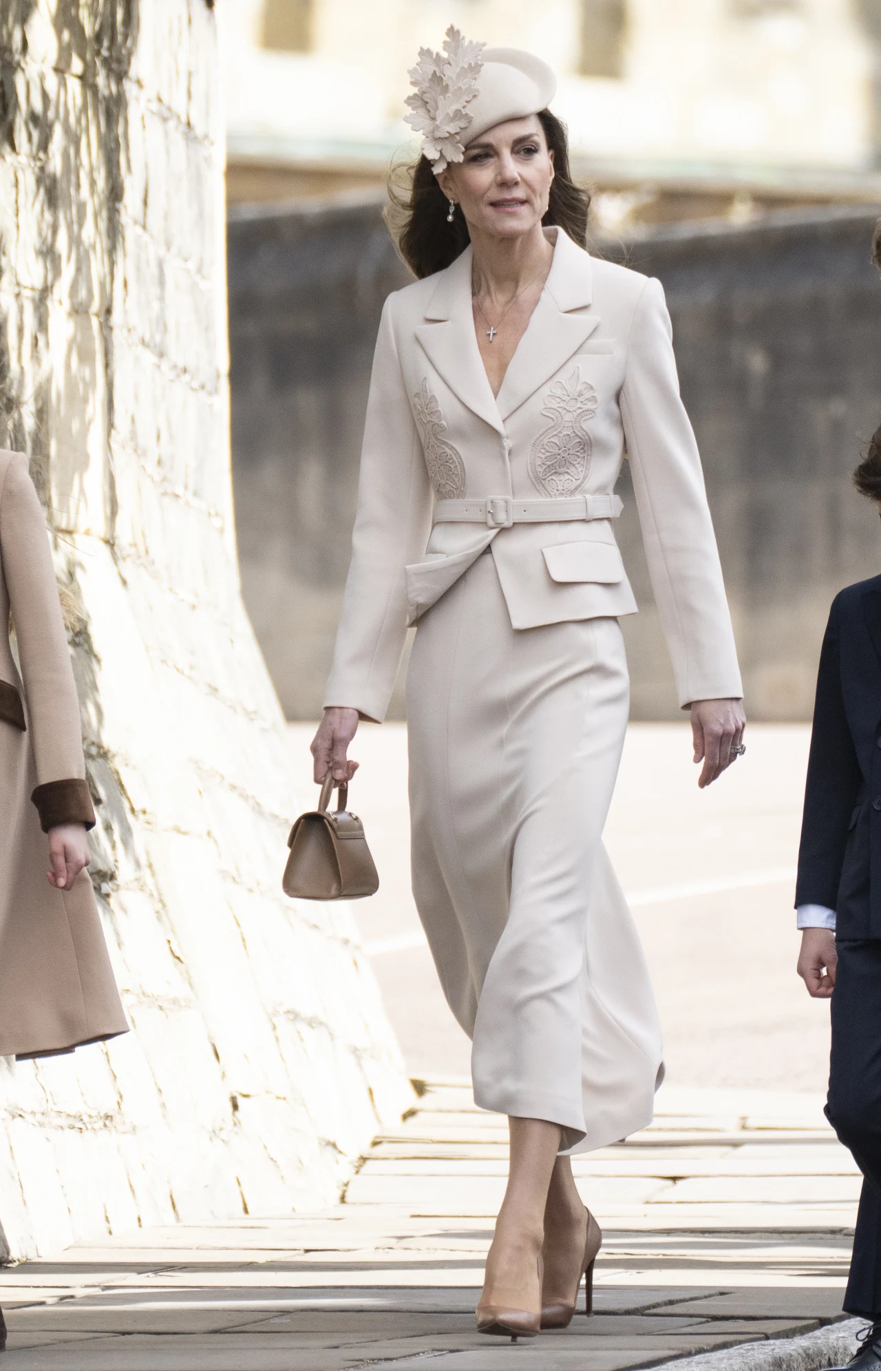 elegancka kobieta w kremowym garniturze z ozdobnymi przeszyciami i spódnicą midi, na głowie ma stylowy kapelusz z dekoracją z liści, w dłoni trzyma małą torebkę w tym samym odcieniu, dopełnieniem kreacji są beżowe szpilki