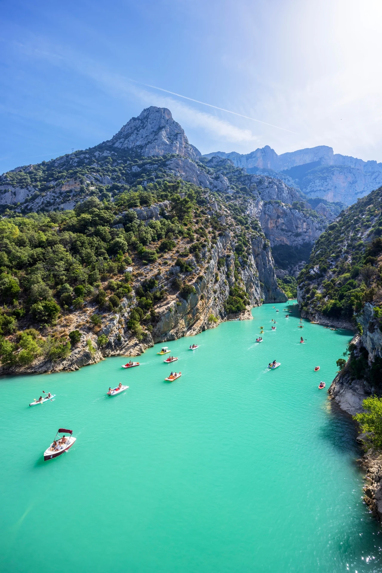 Turkusowa rzeka płynąca przez głęboki wąwóz otoczony stromymi zielonymi zboczami gór, na wodzie liczne kajaki i łódki z wypoczywającymi ludźmi, w tle wysokie, skaliste szczyty pod błękitnym niebem.